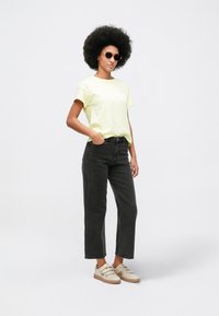 Γυναίκα με αφρικανικό χτένισμα που φοράει γυαλιά ηλίου, ανοιχτό κίτρινο μπλουζάκι, μαύρα τζιν και μπεζ sneakers, στέκεται με το ένα χέρι στην τσέπη.