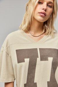 Beige T-shirt met een V-hals ontwerp, voorzien van een grote donkerbruine print aan de voorkant. Zachte stof met een losse pasvorm en korte mouwen.