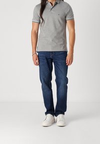 Šedé polokošile s kontrastním lemem na límci v kombinaci s modrými džínami a bílými teniskami. Model stojí s rukama podél těla.