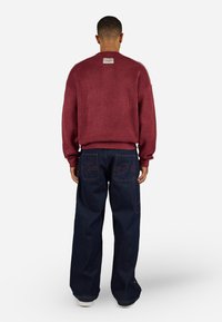 Maroon textured sweater with a ribbed hem and back label, paired with wide-leg dark blue denim pants featuring red stitching and large pockets.