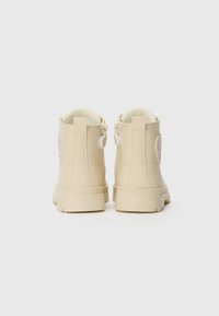 Botas de tobillo beige hechas de material sintético, con una textura suave, cuello acolchado y un parche de corazón colorido en el lado exterior.