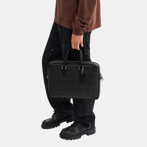 Osoba u smeđoj košulji dugih rukava i crnim hlačama drži crnu torbu s uzorkom za ručke.