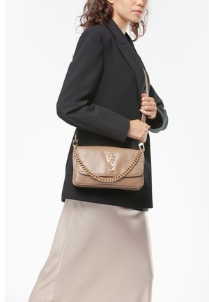 Donna che indossa blazer nero e gonna beige chiaro tiene una borsa taupe con catena dorata e logo sulla tracolla sopra la spalla.