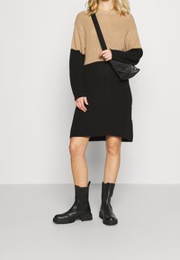 Stickad klänning med beige överdel och svart nederdel. Har långa ärmar, ribbat mönster och sidofickor. Matchas med svarta ankellångstövlar.