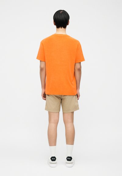 Άτομο που στέκεται με την πλάτη, φοράει πορτοκαλί κοντομάνικο πουκάμισο, μπεζ σορτς, λευκές κάλτσες και λευκά αθλητικά Polo σε απλό φόντο.