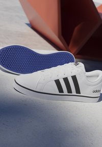 Zapatillas blancas Adidas con rayas negras y suelas azules texturizadas descansando sobre una superficie clara junto a una estructura geométrica de color óxido.