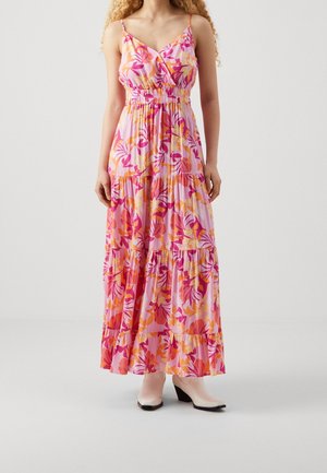 Femme portant une robe maxi rose sans manches avec des motifs floraux orange et fuchsia, associée à des bottines blanches sur un fond uni.