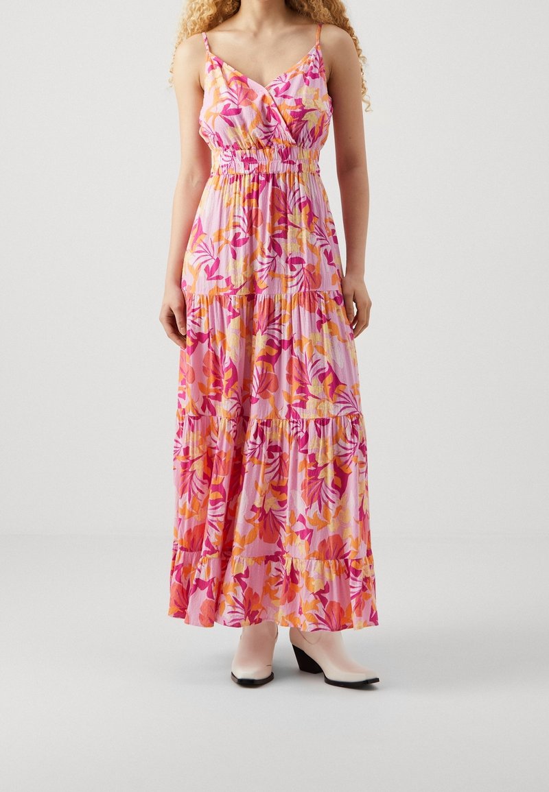 Femme portant une robe maxi rose sans manches avec des motifs floraux orange et fuchsia, associée à des bottines blanches sur un fond uni.