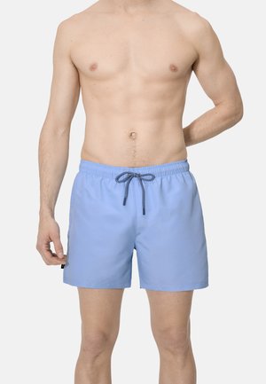 Torso masculino con bañador azul claro con cordón, de pie sobre un fondo liso y claro.