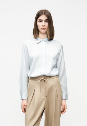 Žena oblečená v svetlomodrej košeli na gombíky, zastrčenej do béžových plisovaných nohavíc, ktoré sú v páse previazané mašľou.