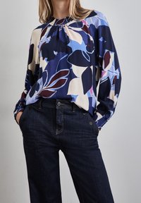 Blusa fluida con un motivo floreale navy, caratterizzata da tonalità di blu, rosa e crema. Collo ed elastico ai polsini, abbinata a jeans in denim scuro.