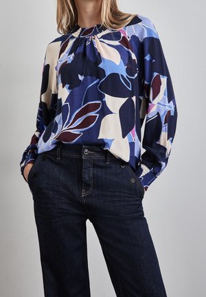 Blusa fluida con un motivo floreale navy, caratterizzata da tonalità di blu, rosa e crema. Collo ed elastico ai polsini, abbinata a jeans in denim scuro.