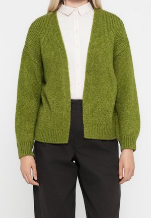 Personne portant une chemise blanche à boutons sous un cardigan ample texturé vert olive à manches longues et un pantalon noir.