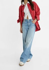 Giacca di jeans rossa sopra una camicia bianca, abbinata a jeans larghi di un blu chiaro. Scarpe casual con una base beige e dettagli bianchi completano l'outfit.