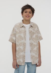 Camicia a maniche corte in tessuto beige con motivi di foglie di palma, design con bottoni e una maglietta bianca sotto. Indossata con jeans azzurri chiari.