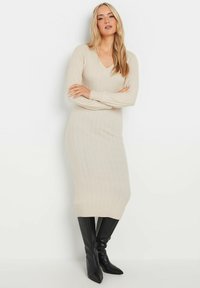 Beiges, geripptes Strickkleid mit V-Ausschnitt und langen Ärmeln, das eine taillierte Silhouette hat und bis zur Mitte der Waden reicht. Stylisch kombiniert mit schwarzen kniehohen Stiefeln.