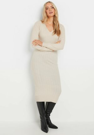 Robe en tricot côtelé beige avec un décolleté en V et des manches longues, présentant une silhouette ajustée et un ourlet qui atteint le mi-mollet. Stylée avec des bottes noires montant jusqu'aux genoux.