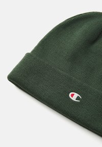 Căciulă tricotată verde cu tiv răsucit și țesătură texturată. Prezintă un mic logo Champion în alb și roșu pe tiv. Material moale și cald.