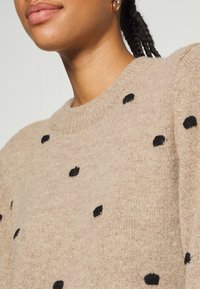 Pull en maille beige avec un col rond, orné de pois noirs, en tissu texturé et épaules tombantes pour une coupe décontractée.