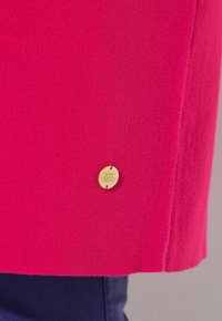 Haut en tricot fuchsia avec une étiquette ronde en métal doré portant l'inscription de la marque. Texture lisse et finition à ourlet droit.