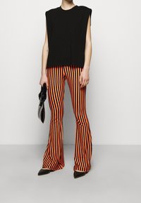 Svart ärmlös topp som kombineras med orange och svarta randiga utsvängda byxor. Modellen håller en svart kuvertväska och bär spetsiga svarta skor.