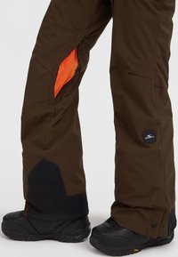Bruine sneeuwbroeken hebben oranje gaasventilatieritsen, zwarte accenten op de onderste boorden en een logo-oppatch aan de zijkant.