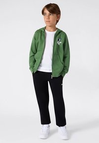 Grüner Kapuzenpullover mit Reißverschluss, Fronttaschen und weißem Cartoon-Motiv, kombiniert mit schwarzen Hosen und weißen Turnschuhen; darunter ein einfaches weißes T-Shirt.