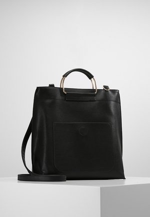 Czarna skórzana torba typu tote z okrągłym złoto-czarnym uchwytem, przednią kieszenią i odpinanym paskiem na ramię na białym piedestale.