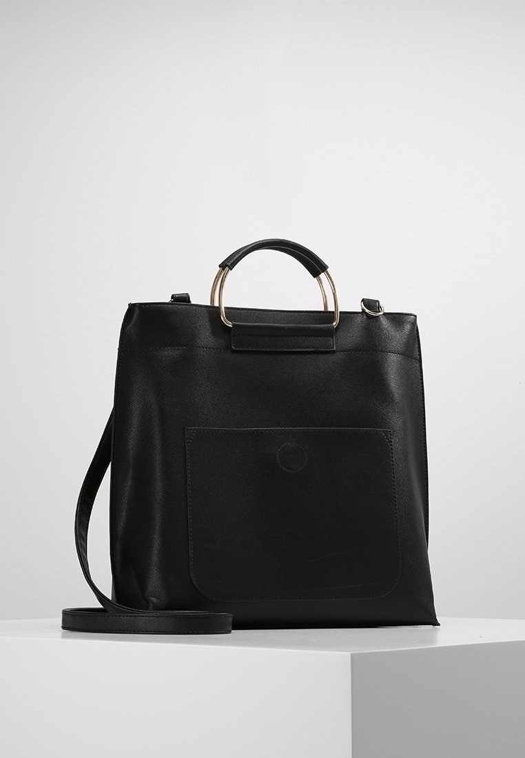 Sac fourre-tout en cuir noir avec poignée ronde en or et noir, poche avant et bandoulière amovible sur un piédestal blanc.