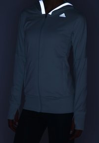 Personne portant un sweat à capuche zippé gris Adidas avec des bandes réfléchissantes sur les bords de la capuche sur un fond sombre.