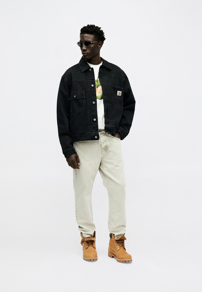 Homme portant une veste en denim noire, un t-shirt blanc à motif, un jean beige clair, des bottes de travail couleur miel et des lunettes de soleil, debout devant un fond uni.