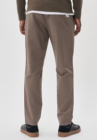 Světlé hnědé kalhoty s hladkou texturou, volného střihu, s délkou po kotníky, nošené s černými teniskami na neutrálním pozadí.