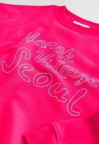 Sweat-shirt rose vif avec texte blanc brodé "Hanok Village Seoul" en écriture cursive sur le devant.