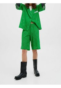 Zielona koronkowa koszula z krótkim rękawem z detalem "1998", zestawiona z dopasowanymi szortami. Czarny botki na obcasie do kostki dopełniają stylizację.