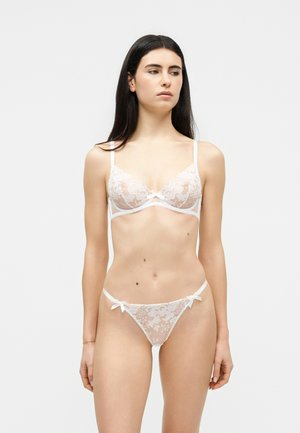 Γυναίκα με μακριά σκούρα μαλλιά, φοράει λευκό δαντελένιο σουτιέν και αντίστοιχο δαντελένιο εσώρουχο, στέκεται μπροστά σε ένα απλό φωτεινό φόντο.