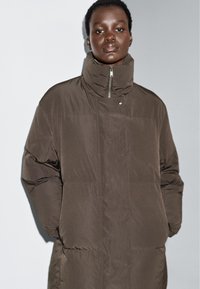 Brun pufferjakke med høj krave, lynlås lukning og store, quiltede sektioner. Har en glat, mat overflade og et minimalistisk design.