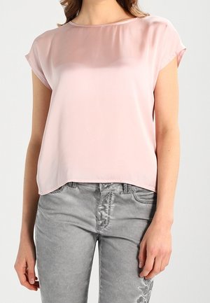 Vrouw draagt een lichtroze doorschijnende blouse met korte mouwen, gecombineerd met grijze jeans met bloemrijke borduursels op het bovenbeen.