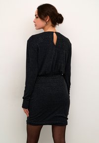 Schwarzes Kleid mit langen Ärmeln, strukturiertem Stoff, taillierter Taille und einem Rückenschlitz. Endet bis zur Mitte des Oberschenkels, kombiniert mit dunklen Strumpfhosen.
