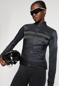 Sort cykeljakke med lynlås foran, reflekterende striber og tekstureret stof. Modellen holder en sort hjelm og bærer solbriller.