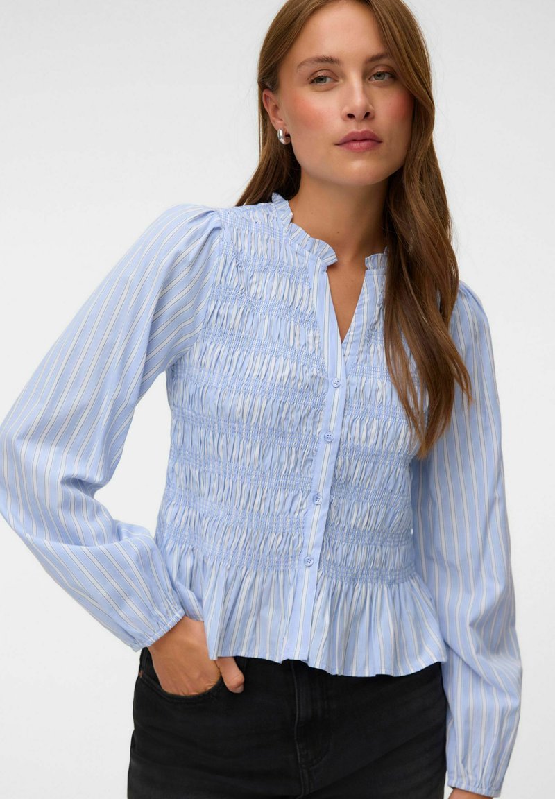 Femme portant un chemisier rayé bleu clair avec devant froncé, fermeture à boutons, ourlet à volants et manches longues bouffantes, associé à un pantalon noir.