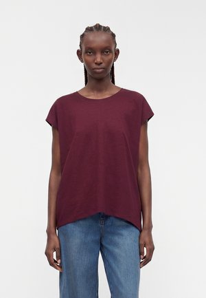 Femme aux cheveux tressés portant un haut ample bordeaux à manches courtes et un jean bleu, debout devant un fond clair uni.