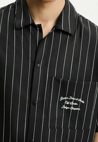 Černá košile na knoflíky s bílými vertikálními pruhy, s náprsní kapsou a vyšitým textem. Hladká tkanina s klasickým designem límečku.