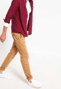 Burgundyröd långärmad skjorta över en vit t-shirt, kombinerad med beige chinos och vita sneakers, som visar en avslappnad passform och uppvikta ärmmanschetter.