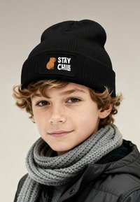 Black knit beanie with a "Stay Chill" patch featuring a bear illustration; worn with a gray knitted scarf and black jacket.