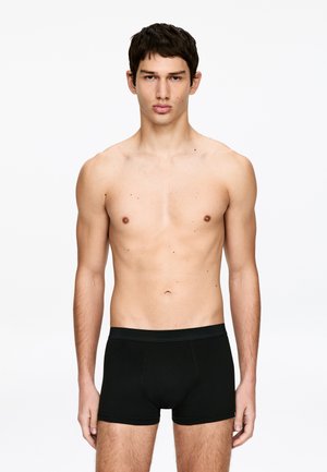 Jeune homme aux cheveux courts et foncés, portant un boxer noir, debout sur un fond blanc uni.