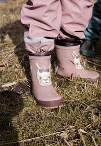 Bottes en caoutchouc roses avec un motif de lama souriant, dotées d'une bordure brune et d'une semelle texturée, posées sur un sol herbeux couvert de paille.