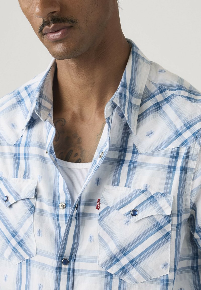 Mand iført blå og hvide ternet skjorte over en hvid ribstrikket tanktop, med brysttatovering delvist synlig.