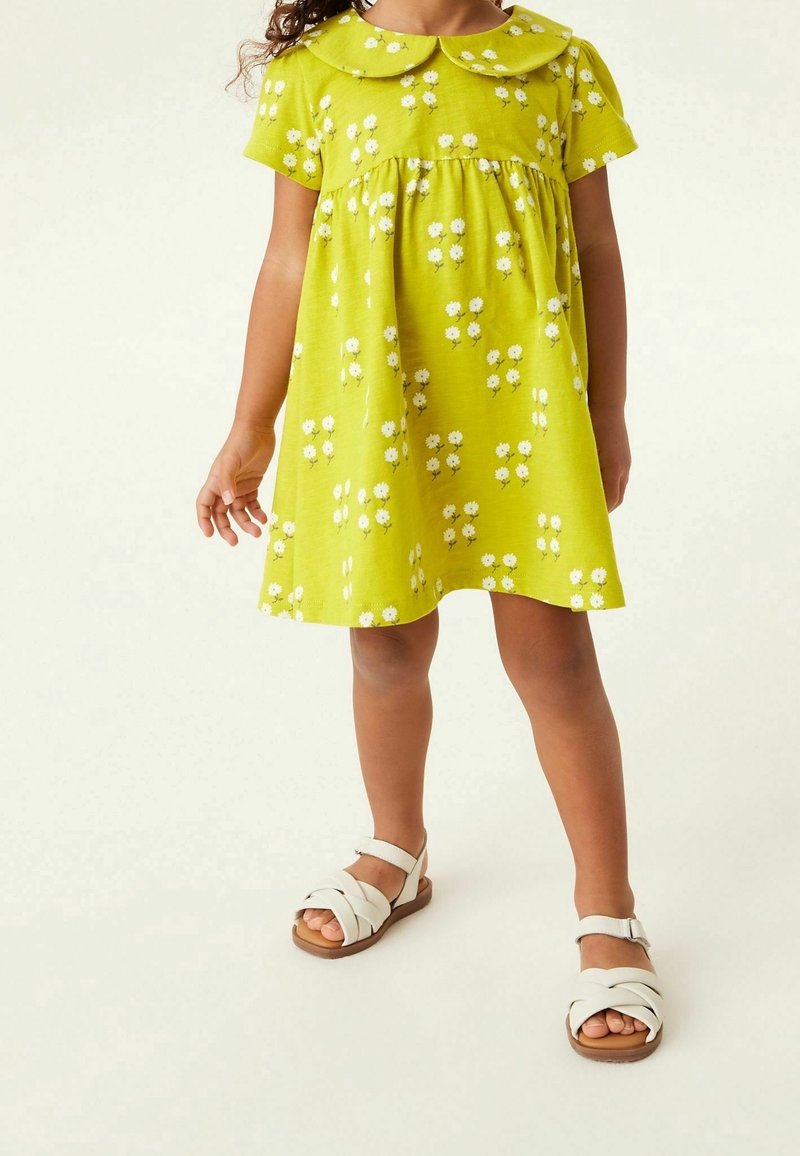Bambina che indossa un vestito giallo con motivi floreali bianchi e sandali bianchi, in piedi su uno sfondo chiaro e uniforme.