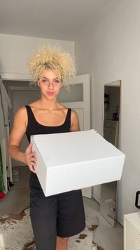 Una scatola di cartone bianco tenuta da una persona con una canottiera nera e pantaloni corti. La scatola ha delle alette sopra, posizionata in un interno luminoso e chiaro.