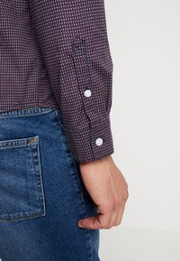Camicia a maniche lunghe di colore rosso scuro con motivo geometrico bianco e blu, dotata di polsini con bottoni e tessuto testurizzato, abbinata a jeans blu.